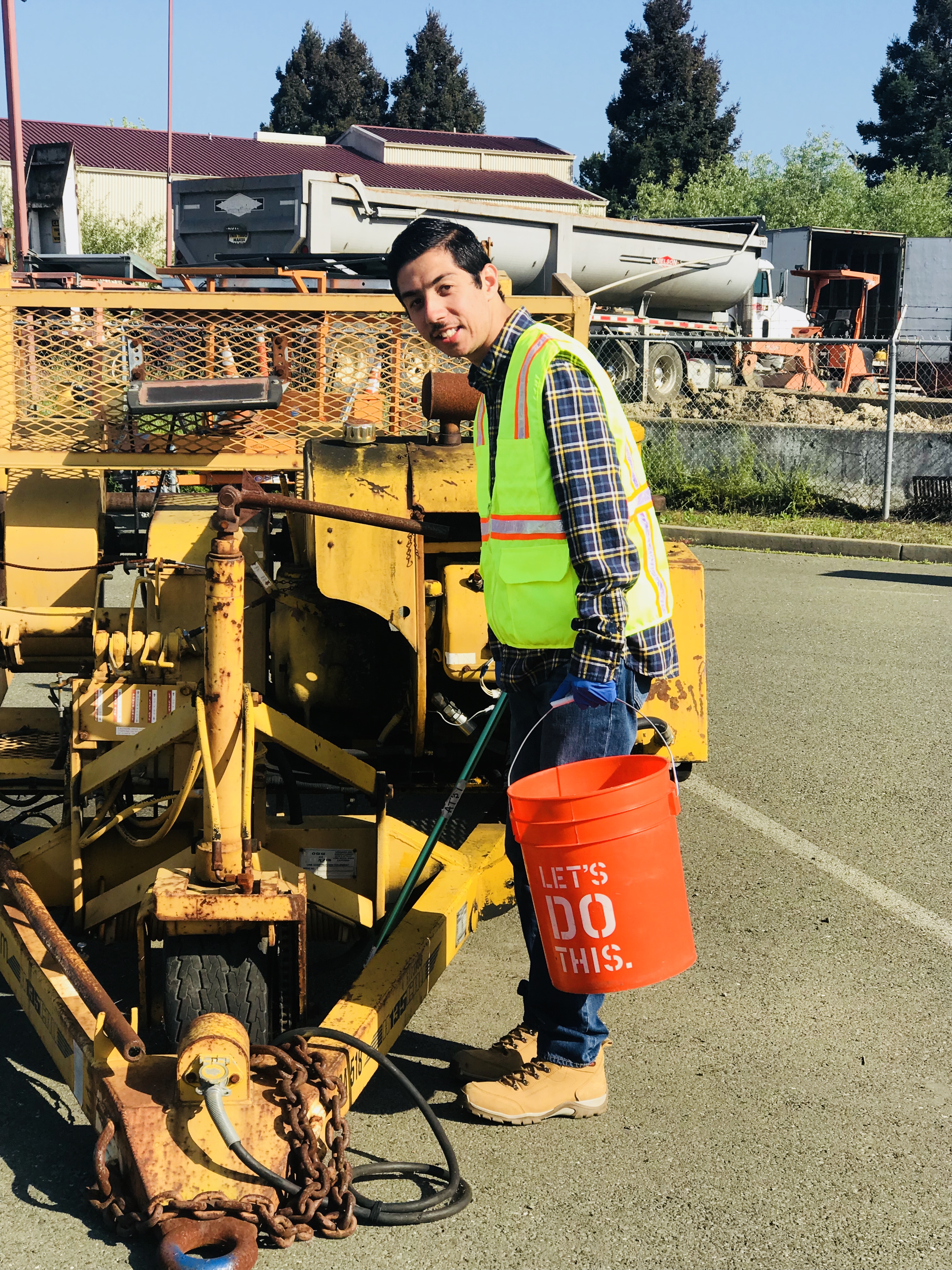 young man in a safety vest holding an orange bucket while standing next to heavy equipment